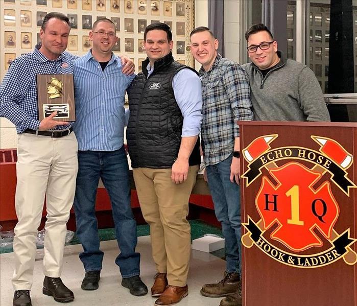 Servpro employee holding an awarf plaque with 4 firemen